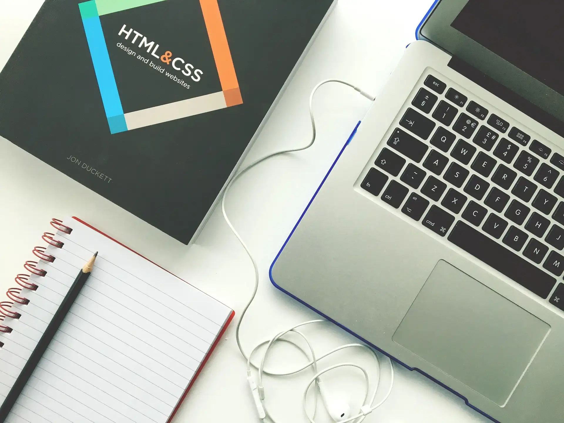 HTML and CSS design book next to laptop, notebook, pen, mouse, and earbuds on white desk