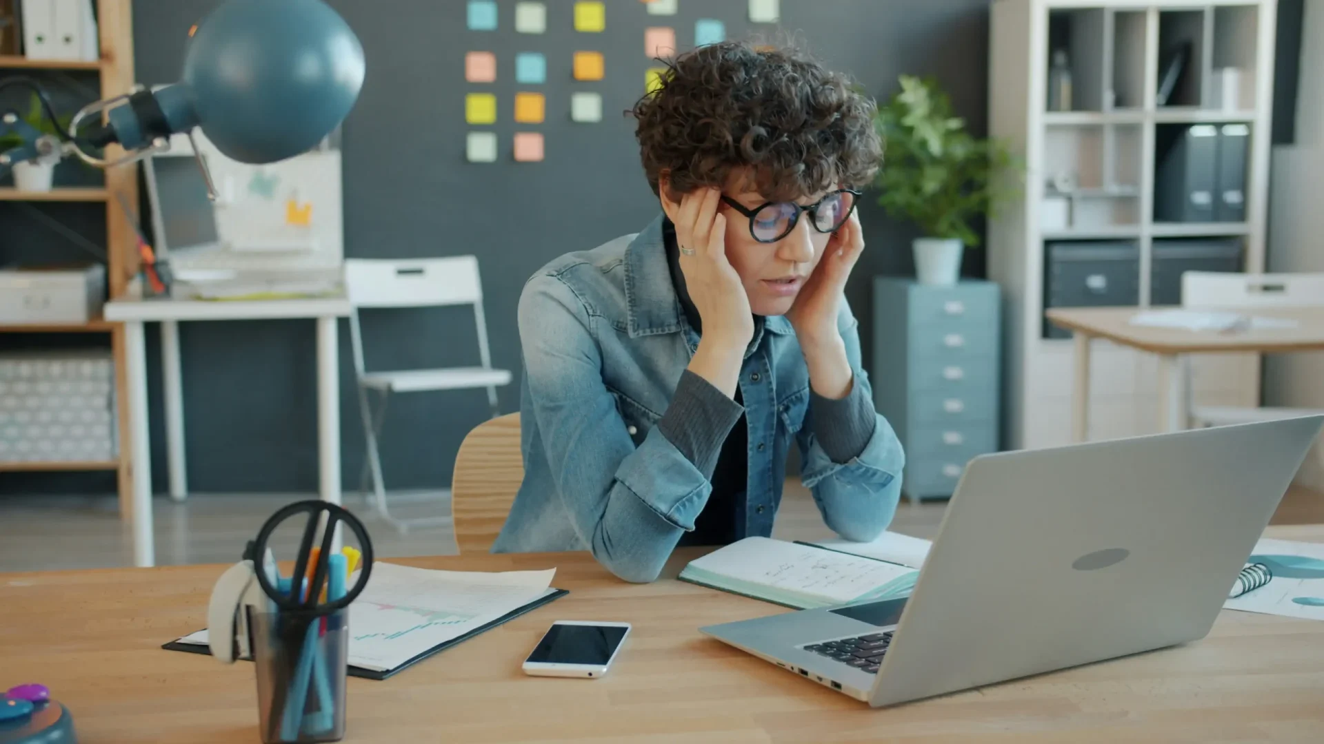Person in denim shirt with glasses looking stressed at desk with laptop